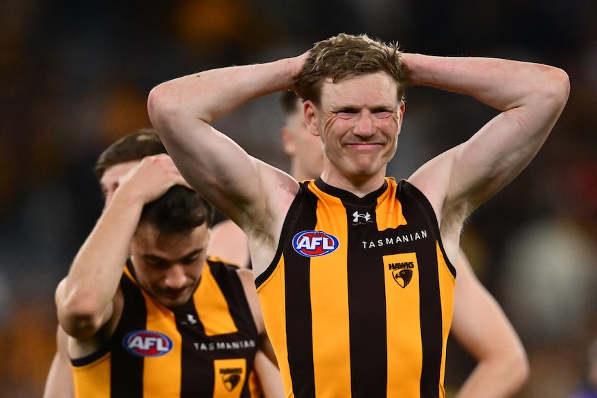 A dark-haired AFL player grimaces while holding his hands to his head during a match.