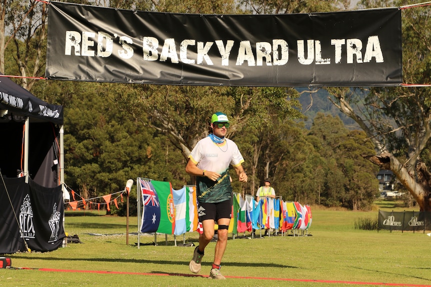 Dan runs underneath a finishing sign wearing a white and green shirt, black shorts and a green hat.