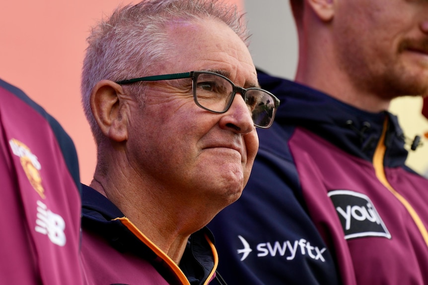 A man with short grey hair and dark rimmed glasses stands in a line with other men in maroon and blue polo shirts.