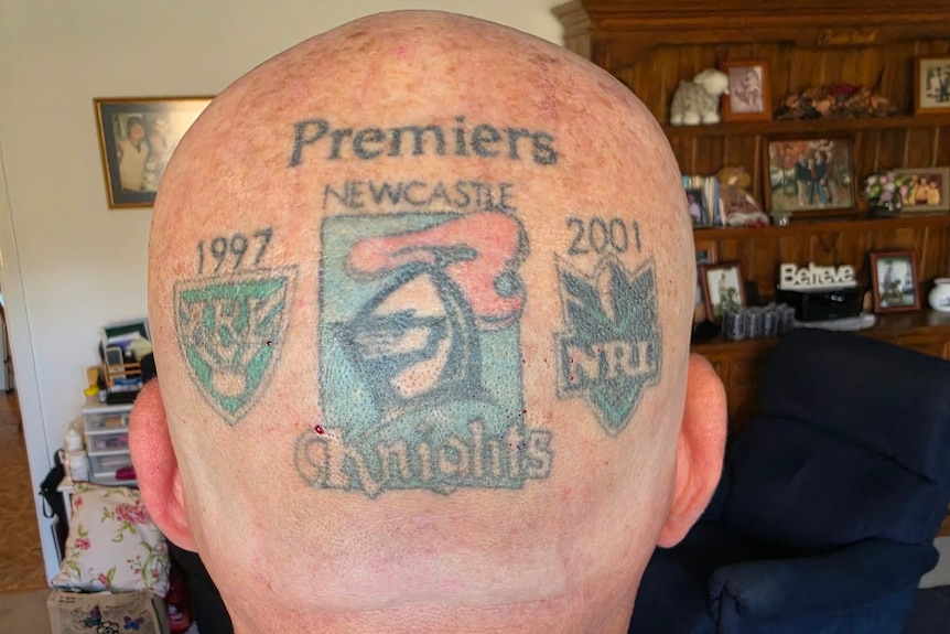 The back of a bald head with three tattoos - a knights emblem, and NRL premiership shields for 97 and 2001.