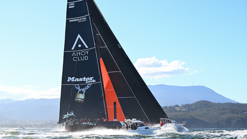 A supermaxi yacht sails up the Derwent River towards Hobart with small craft around it.