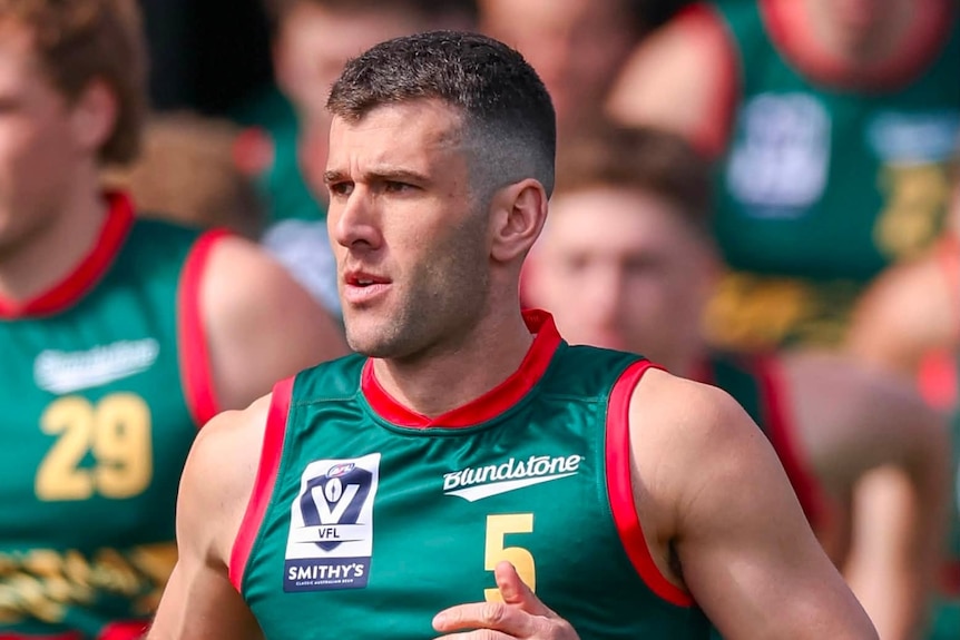Robbie Fox wears a Tasmania Devils football guernsey and stands in front of some teammates.
