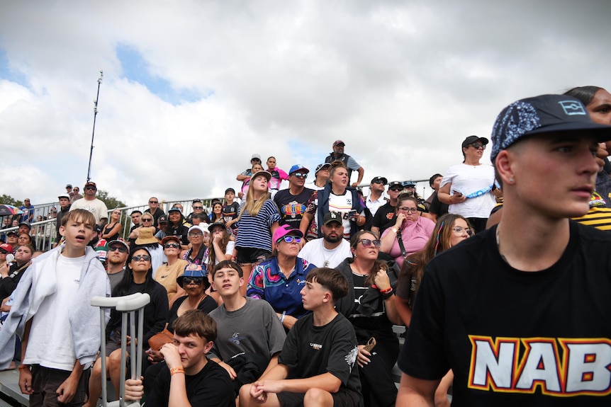 A group of men and women in a grand stand look over the camera.