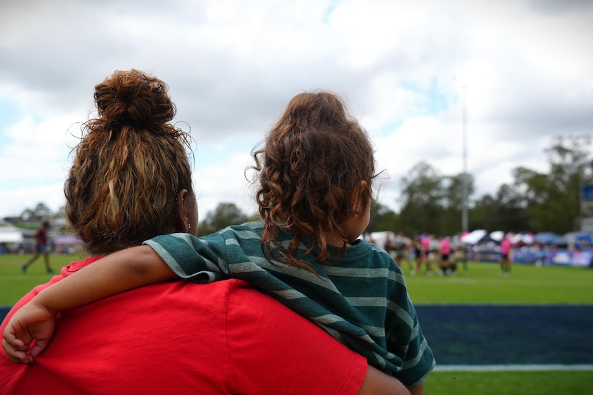 A woman with curly hair holds a young girl, backs to camera. They are watching rugby game on a grassy field.